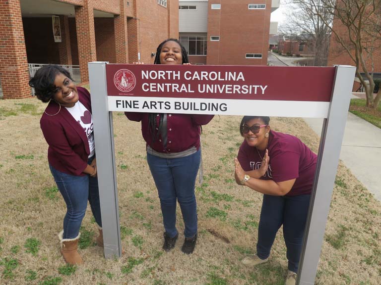 3 students posing by the Fine Arts Building sign