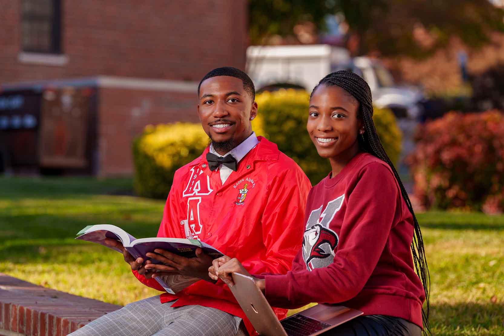 Two students sitting on a low wall outside smiling at the camera