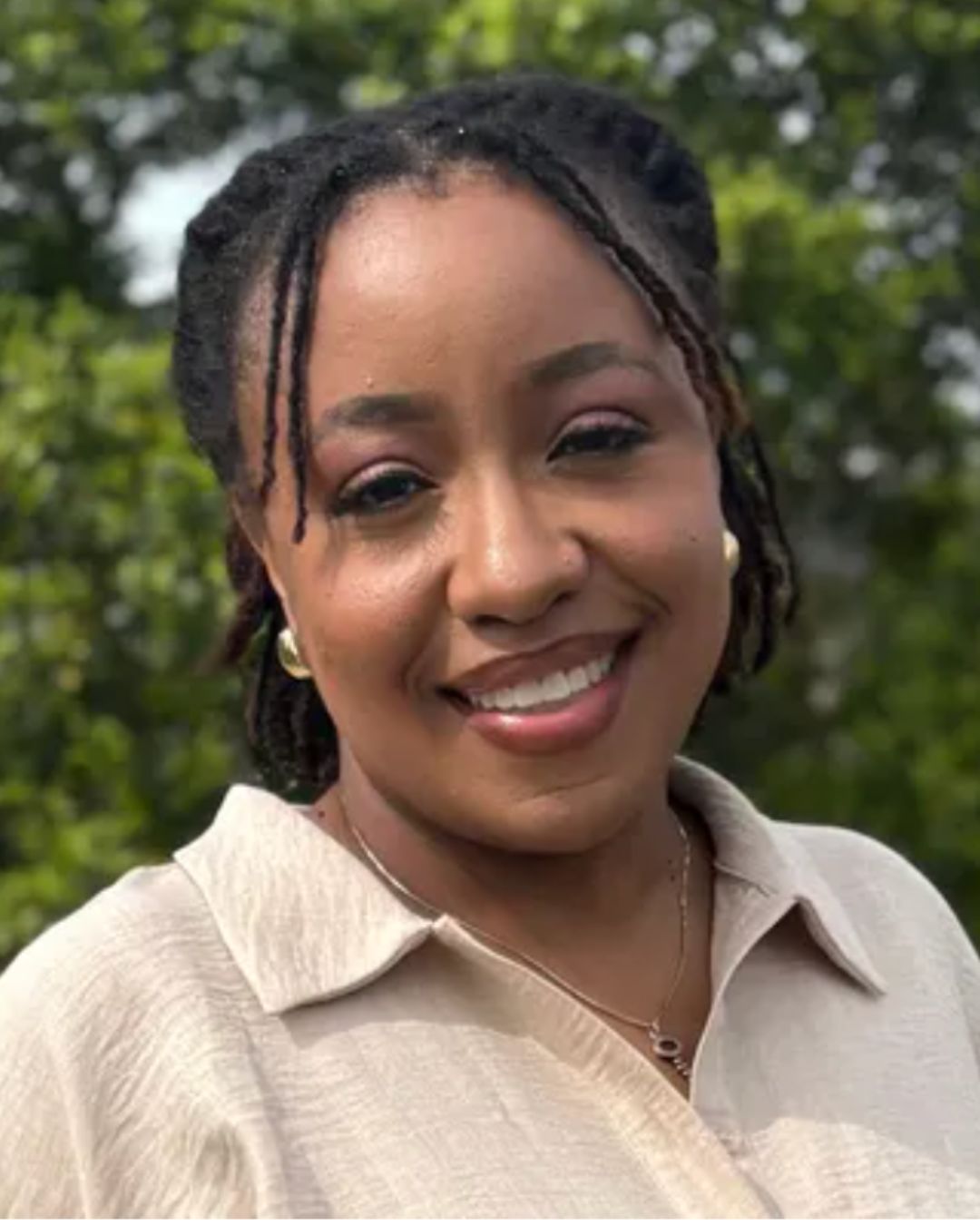 Smiling woman outdoors in a beige shirt with greenery behind her.