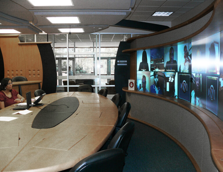 a person sitting alone at a conference table looking at a wall of telepresence screens. 