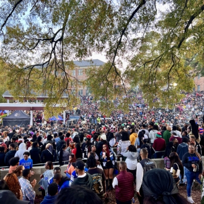 A large crowd gathered outdoors under tree branches at a campus event, with people standing shoulder to shoulder and a brick building visible in the background.