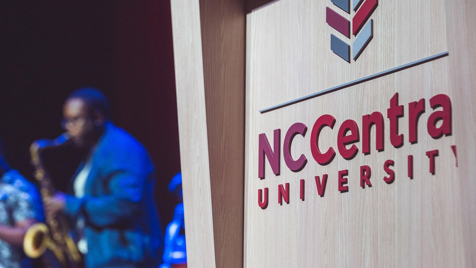 The image shows the "NCCentral University" logo on a wooden surface, with a person playing a saxophone in the blurred background.