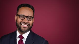 Man wearing suit and glasses smiling on a maroon background.