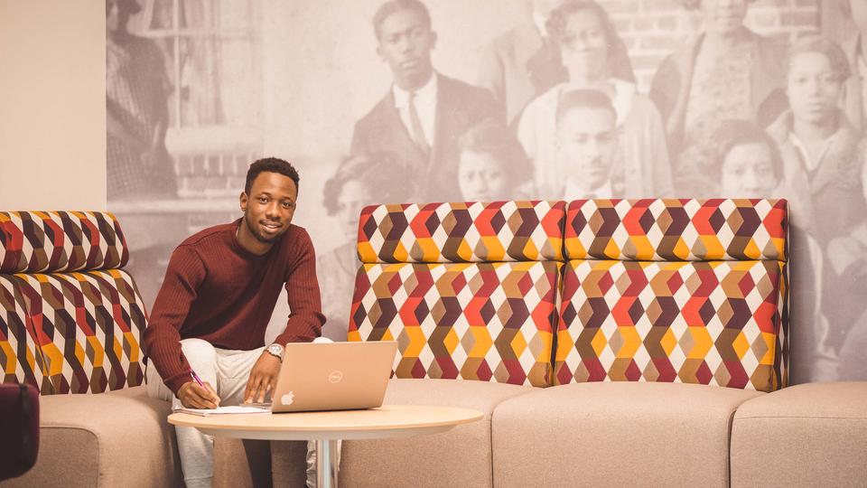 A man with a laptop looking at the camera as he poses in a lounge area