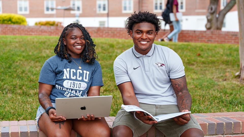NCCU students sitting down together. One holding a laptop and one holding a book